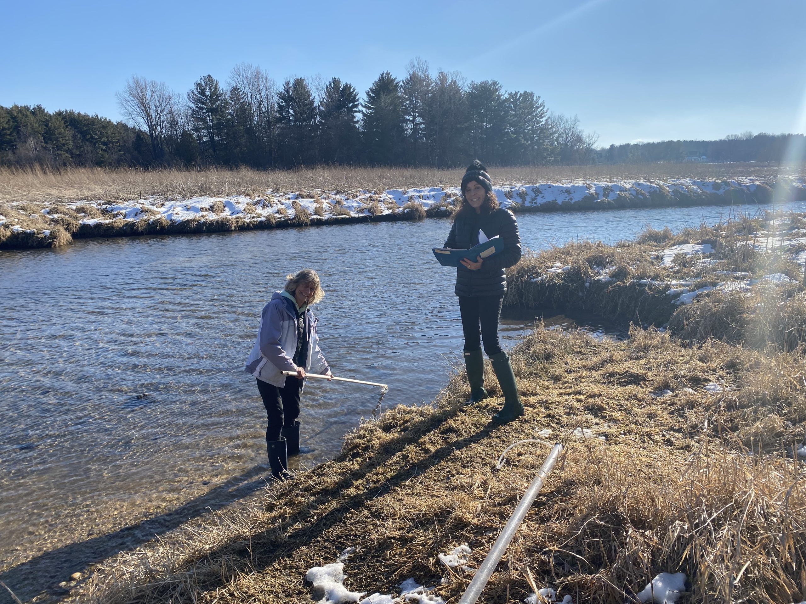 Two volunteers smile at camera standing alongside a snowy creek on a sunny day.