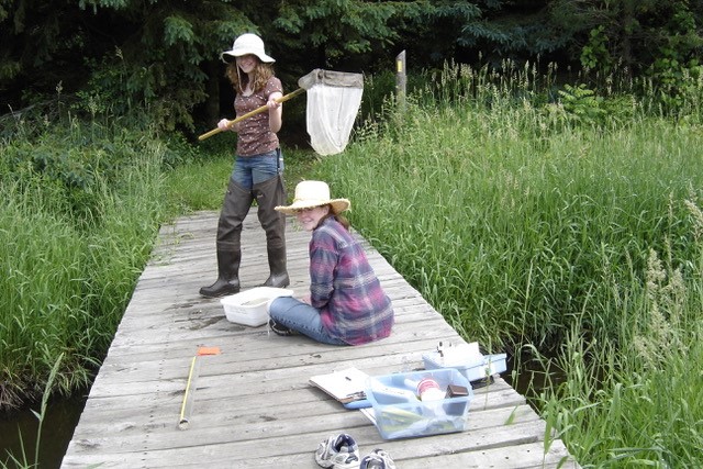 Two women are on dock wearing bucket hats. One is standing wearing waders and holding a net. The other sits on the dock with a white tray in front of her. 