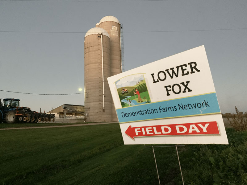 A sign saying lower fox demonstration farms network field day in the foreground with a red arrow pointing to a farm in the background