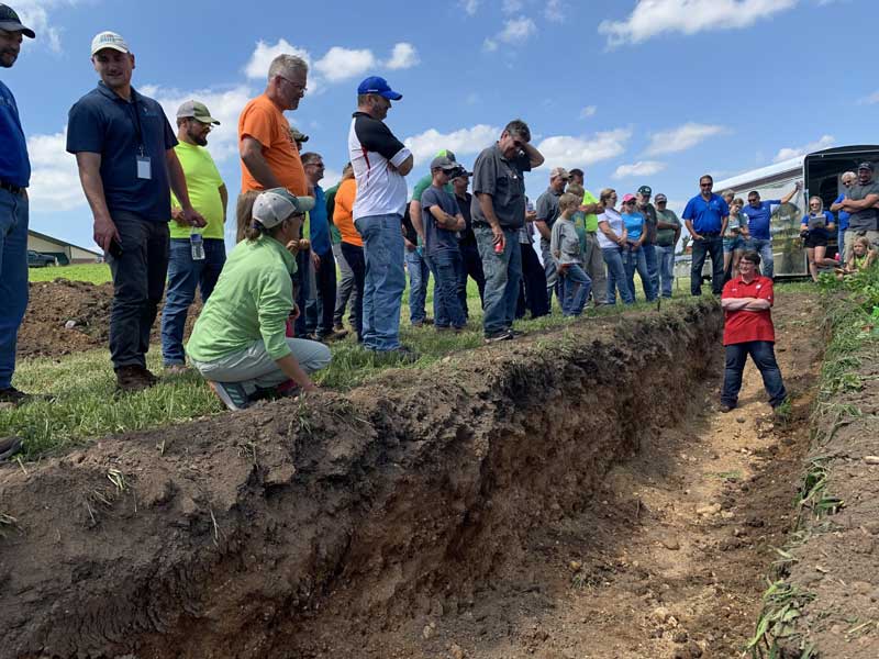 A group of people standing on the edge of a trench while a man inside the trench speaks to them