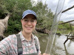 A woman standing in front of a river holding a large glass tube