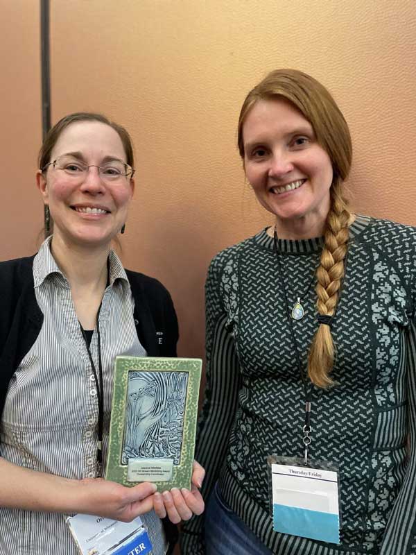 Two women standing beside one another. One is holding an award