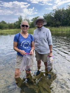 Two women standing in a river holding clear plastic bags with shells in them