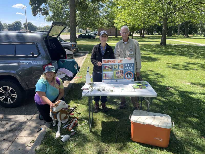 Three people and a dog standing in front of a table holding a snapshot day sign in a park near a lake
