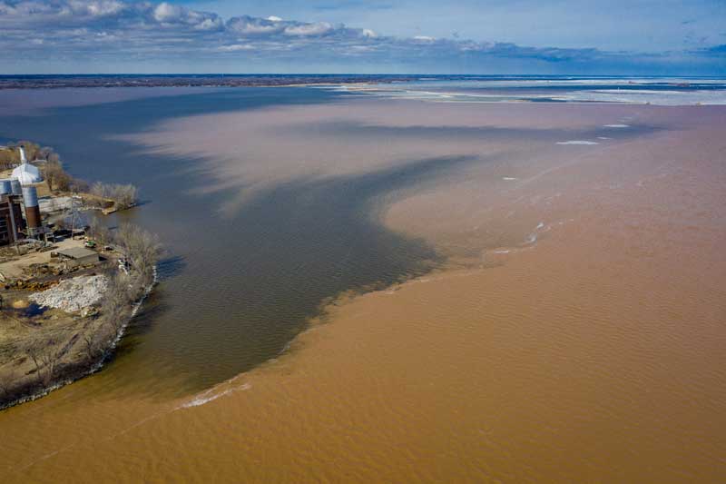 An aerial view of a farm that sits close to the shoreline of a large brown body of water with large dark areas of water around the shoreline.