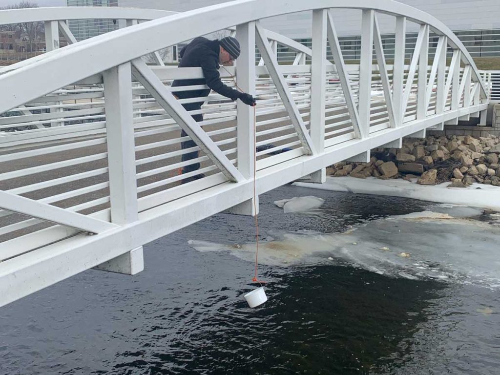 A man dangling a bucket on a string over a semi frozen river from a white bridge in the winter