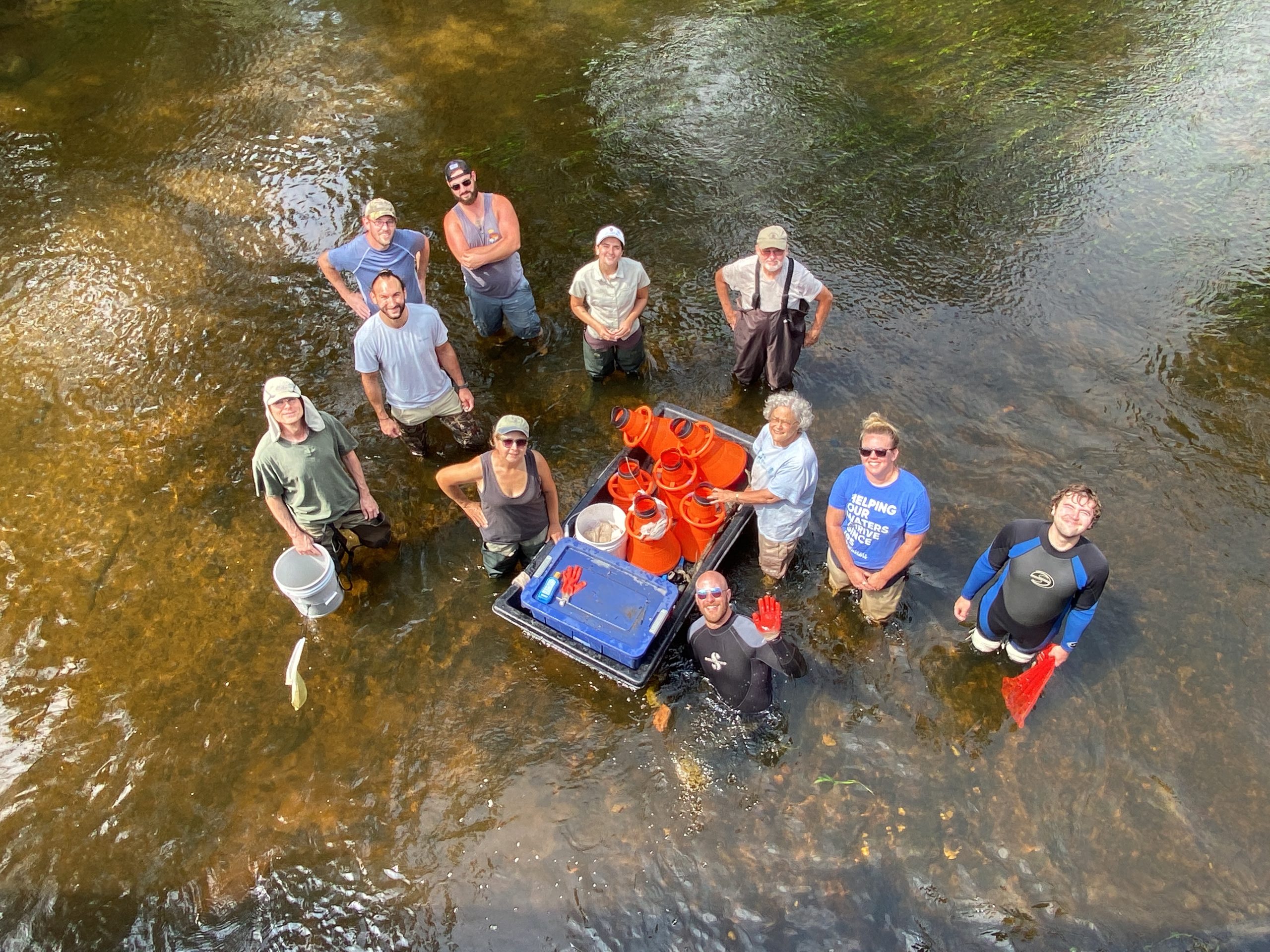An overhead camera shot, with a group of people standing in a shallow river, smiling up at the camera. There is a floating container holding many buckets and supplies.