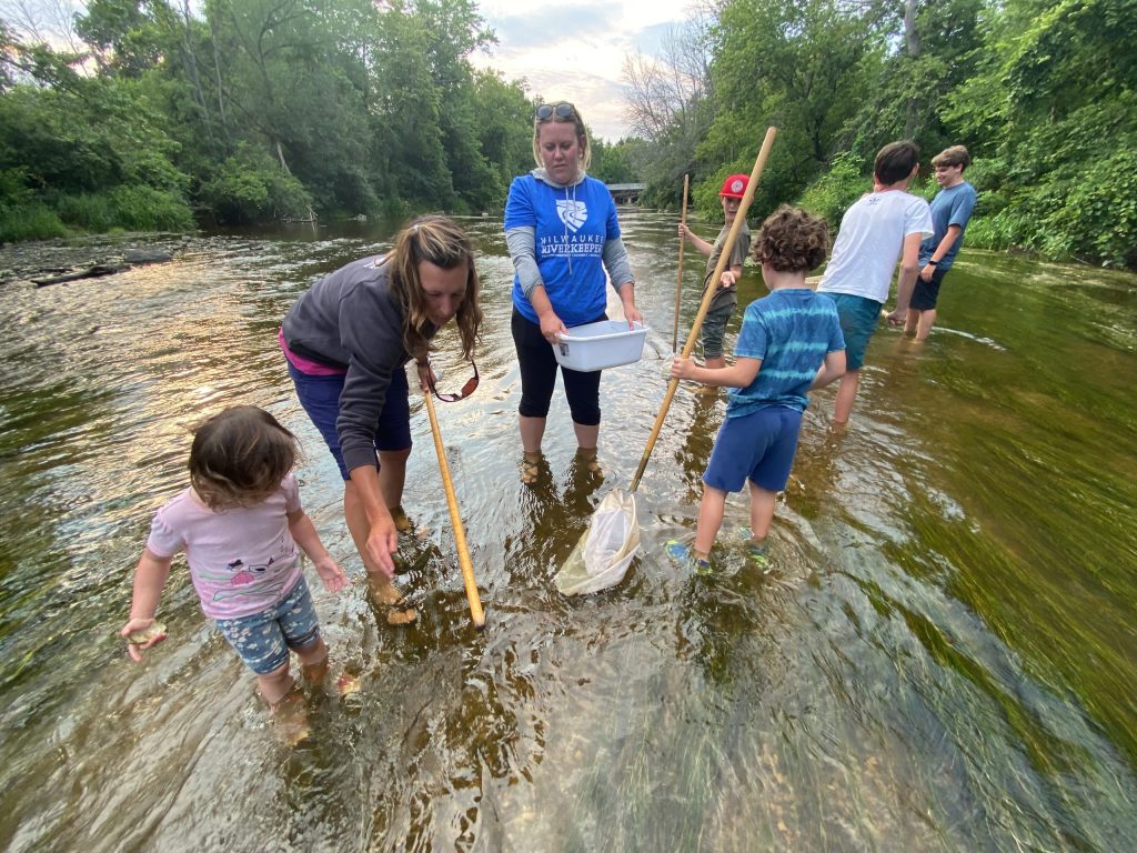 Two adults, including Katie, stand in a shallow stream and help a group of children collect aquatic bugs with nets. Katie is holding a white bin.