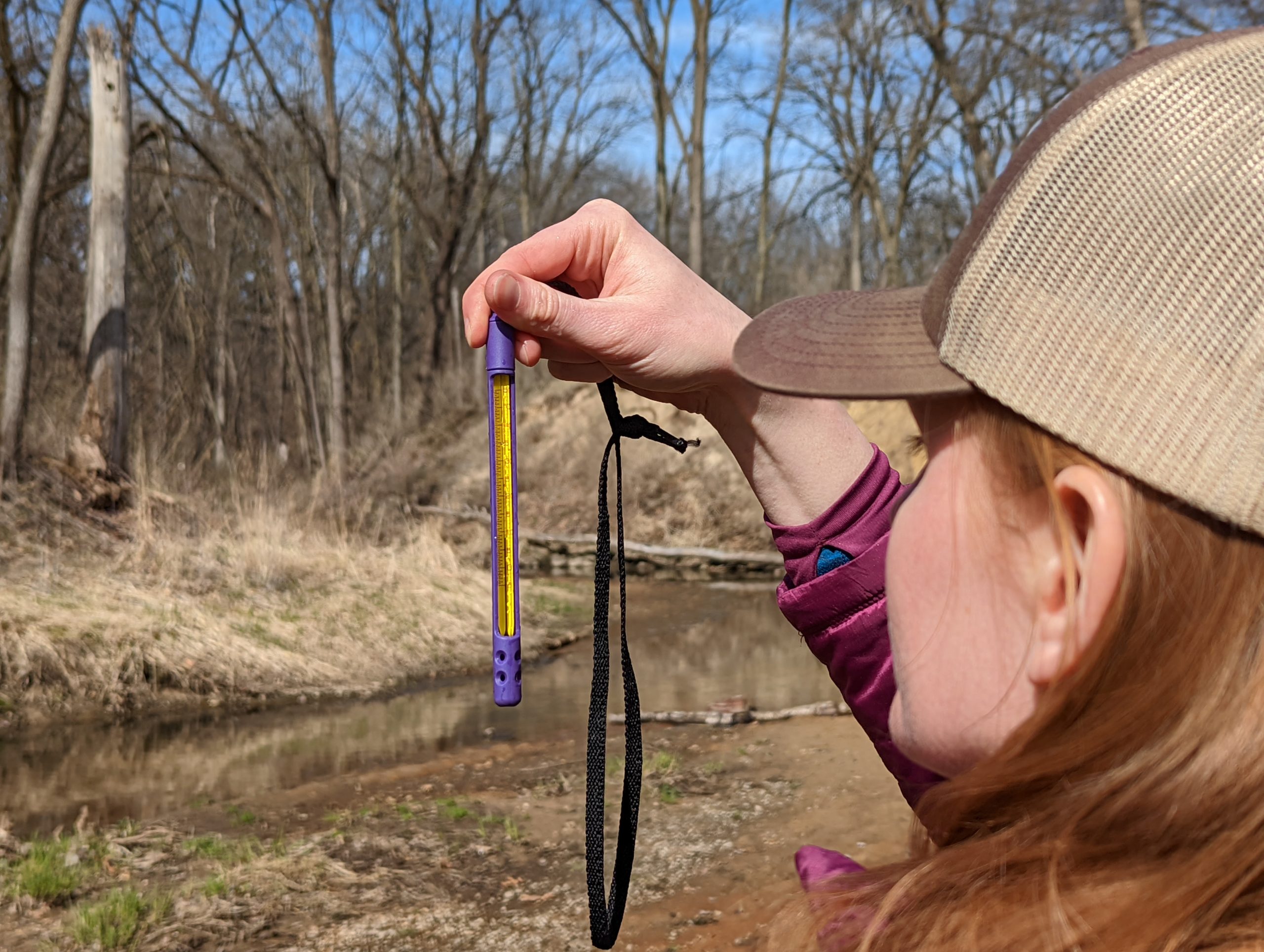 A person holds a thermometer at eye level next to a stream.