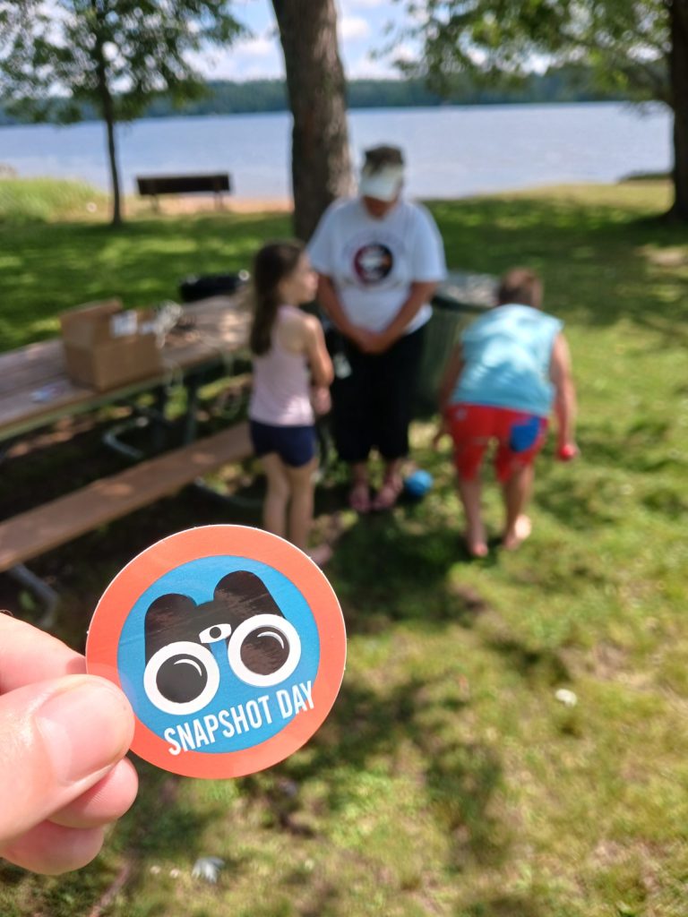 A sticker of the Snapshot Day logo (a blue circle with an orange border and black binoculars in the center). In the background are two young volunteer and an adult volunteer sitting on a bench. 