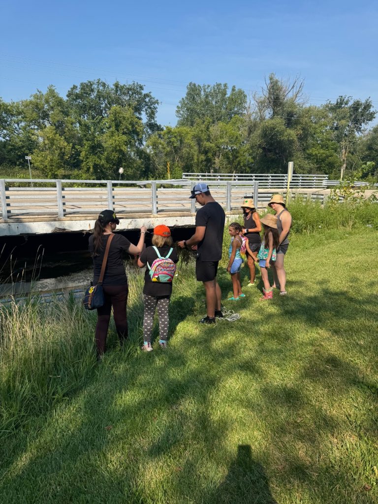 A group of seven people, adults and children, stand looking a stream. It is a sunny day. 