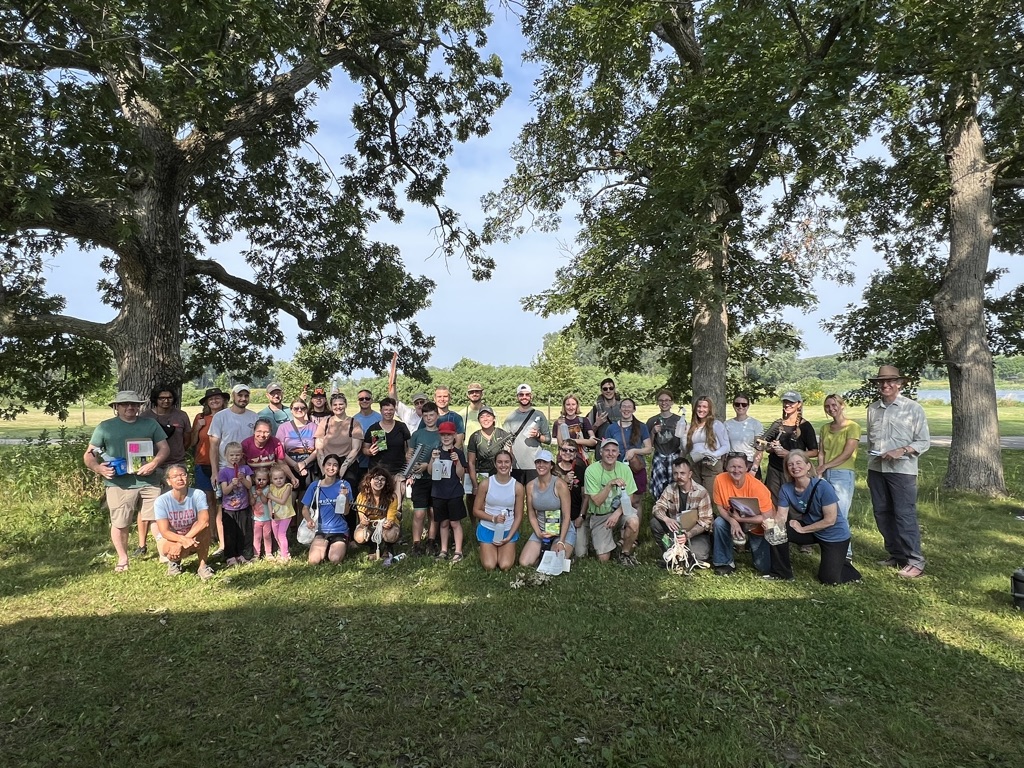 A group of volunteers stand posing for a photo, smiling. It is a sunny day and they are standing between two trees. 