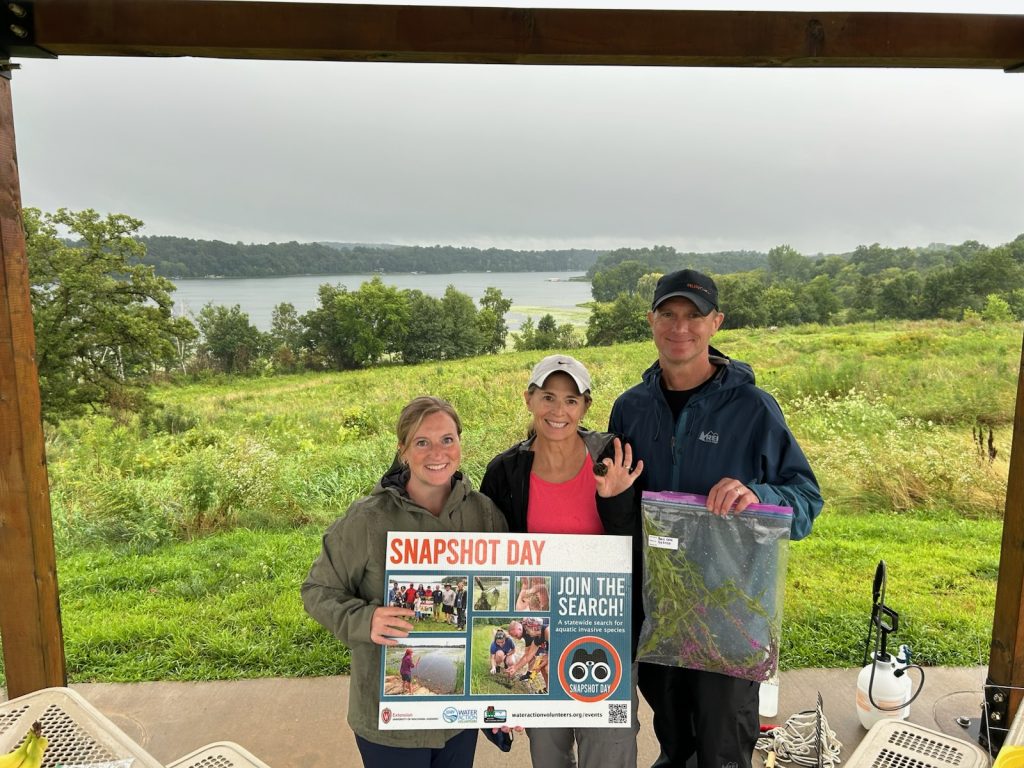 Three volunteers are smiling holding a yard sign and a Ziploc bag full of invasive aquatic plants. The background is of a river. 