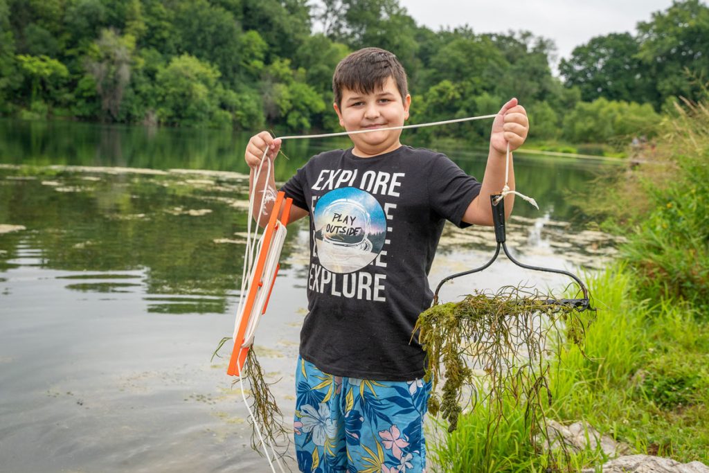 A boy holding a rake head tied to a rope. The rake is full of weeds retrieved from the stream behind him.