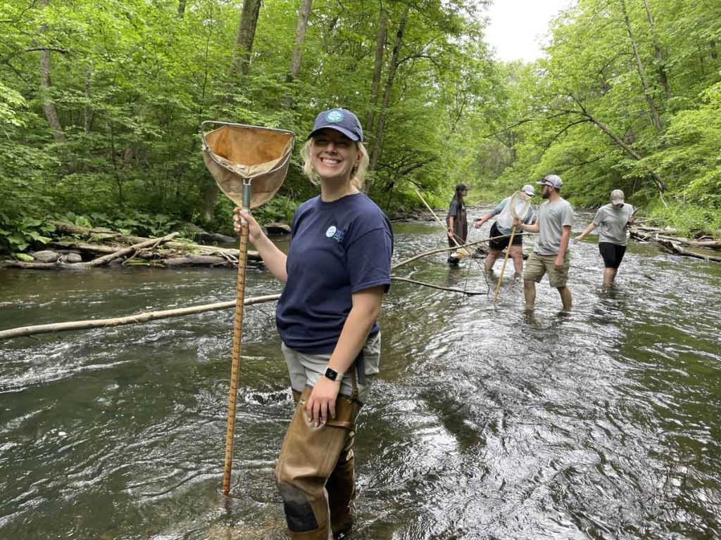 A woman standing in a stream in front of a small group of people. They are using nets to capture invasive species in the stream.