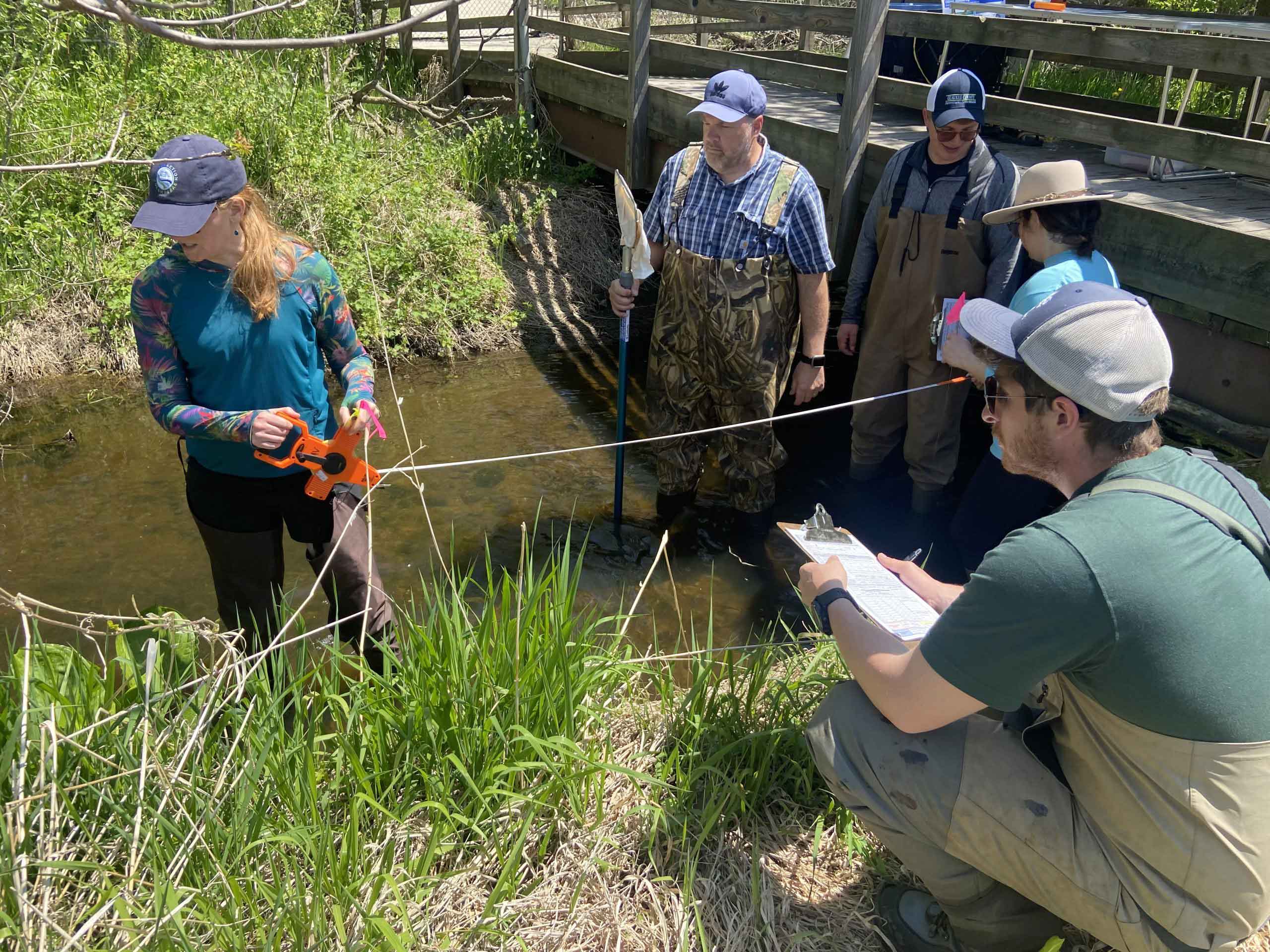 A small group of people measuring a stream's bank near a bridge.