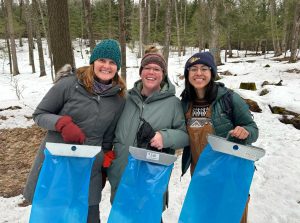 The three WAV staff smile with their blue maple syrup collection bags in a snowy forest.