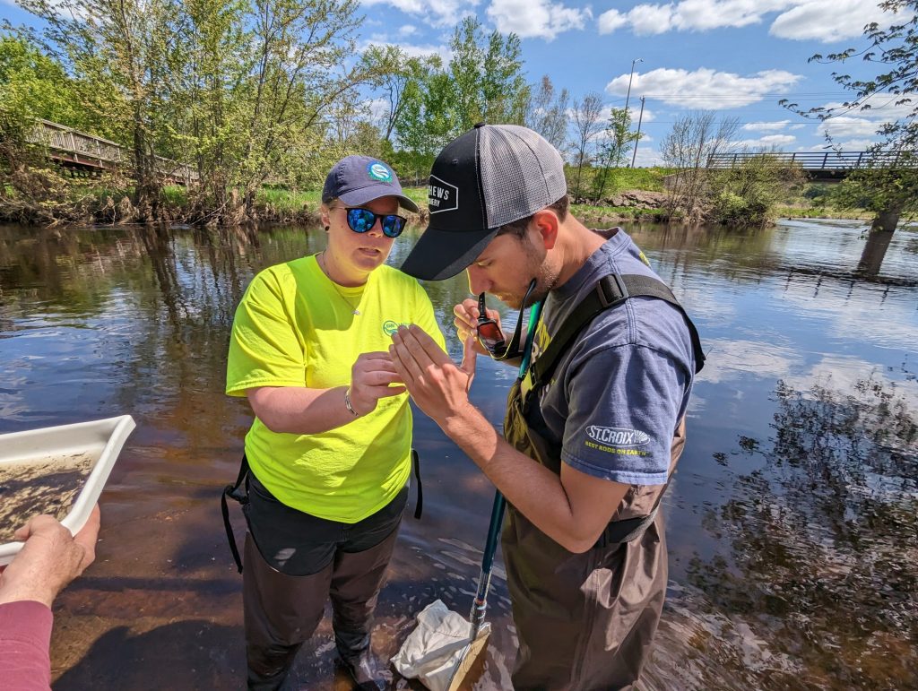Two people stand on the shore of a river looking at a macroinvertebrate.