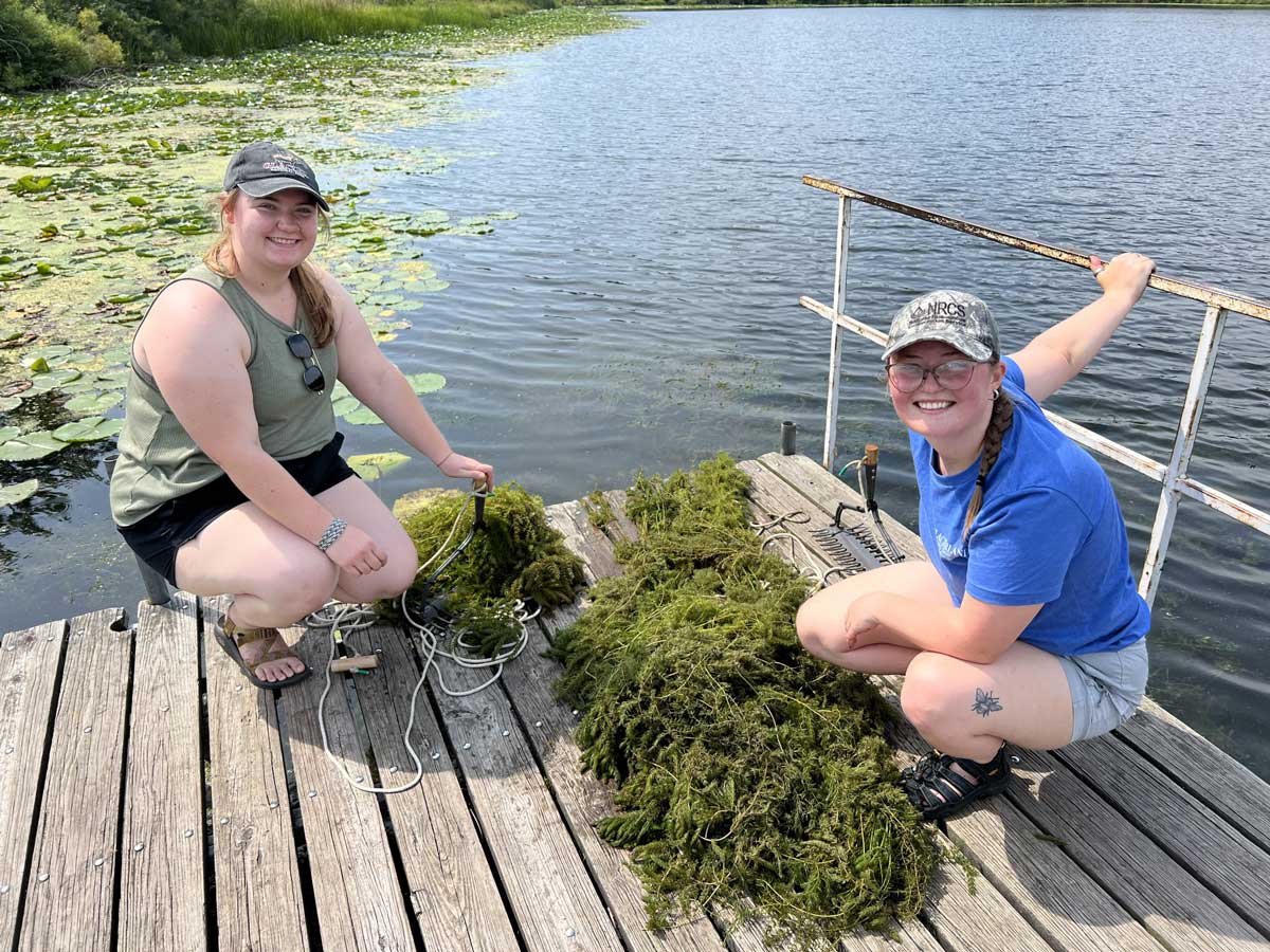 Two girls on a dock removing aquatic plants from a lake