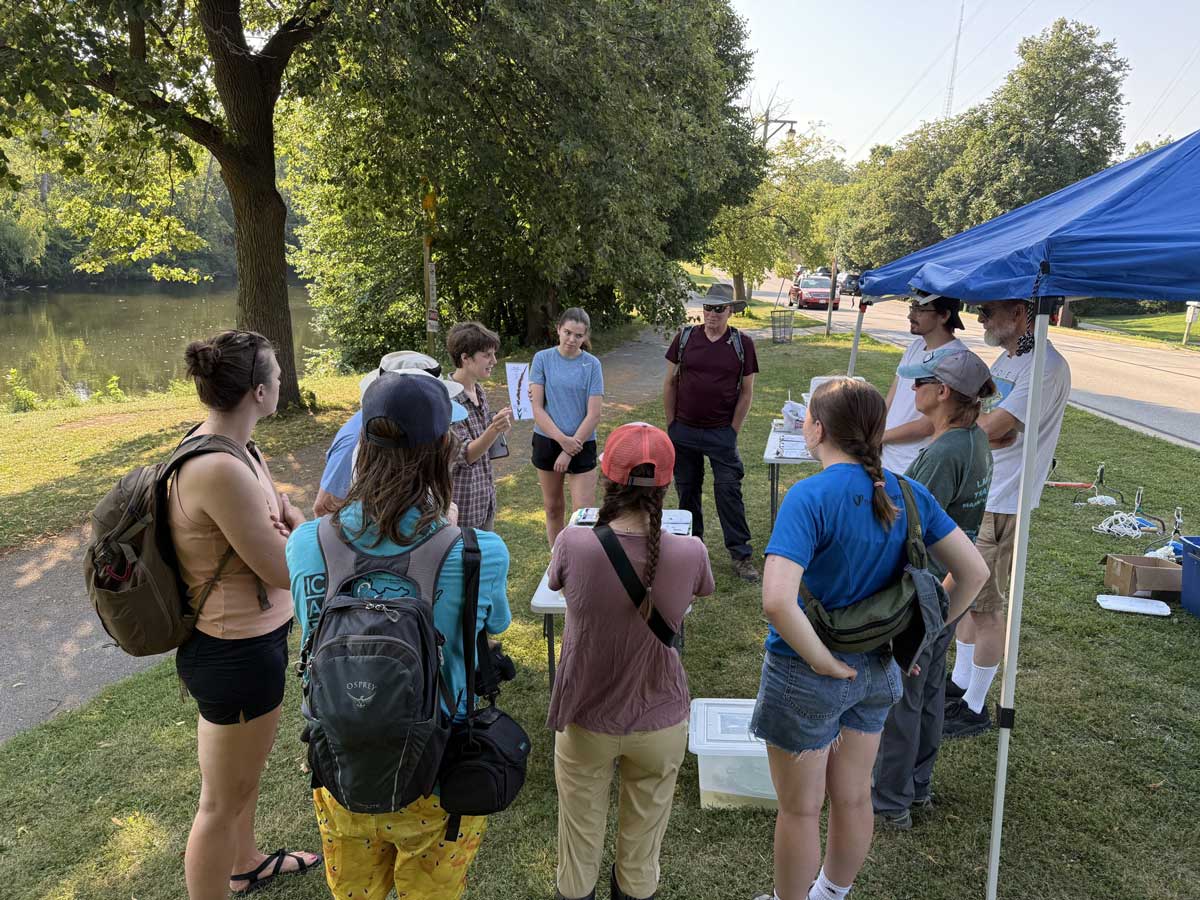 A large group of young people in a park area listening to a speaker
