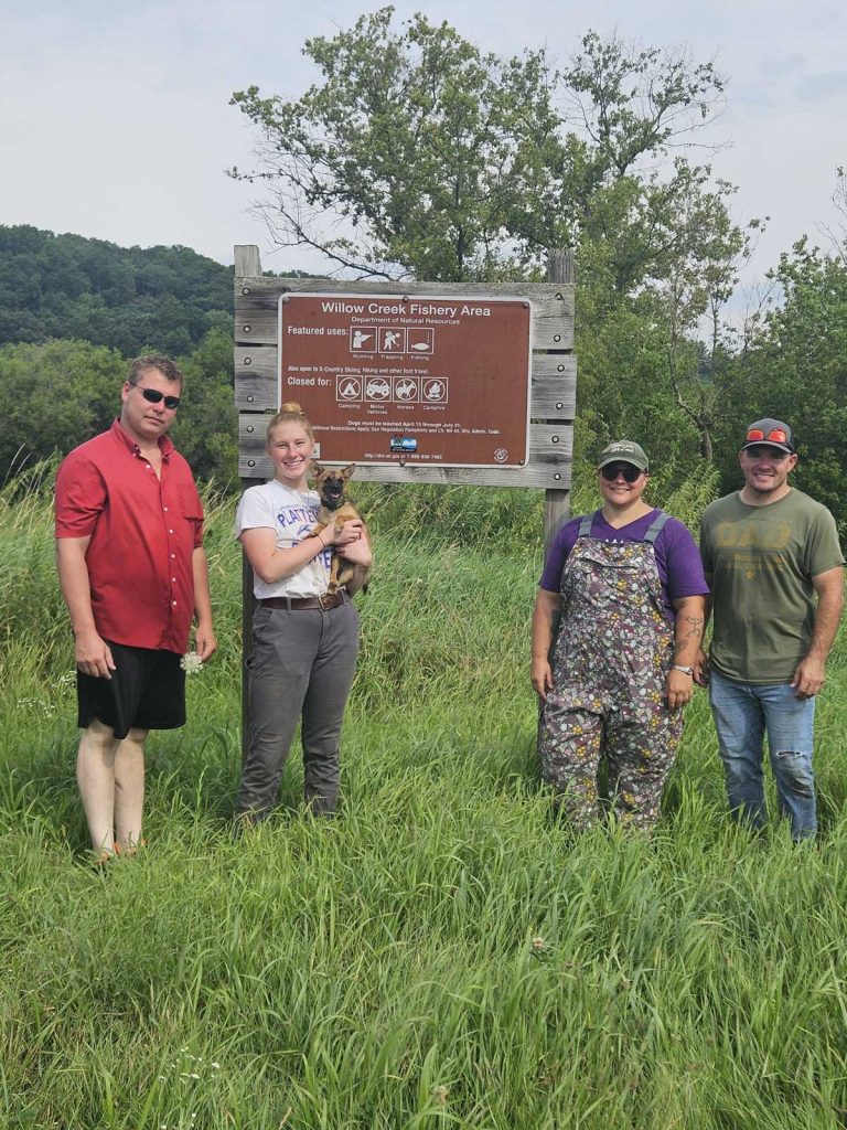 Four people standing in a field in front of a Willow Creek Fishery Area sign