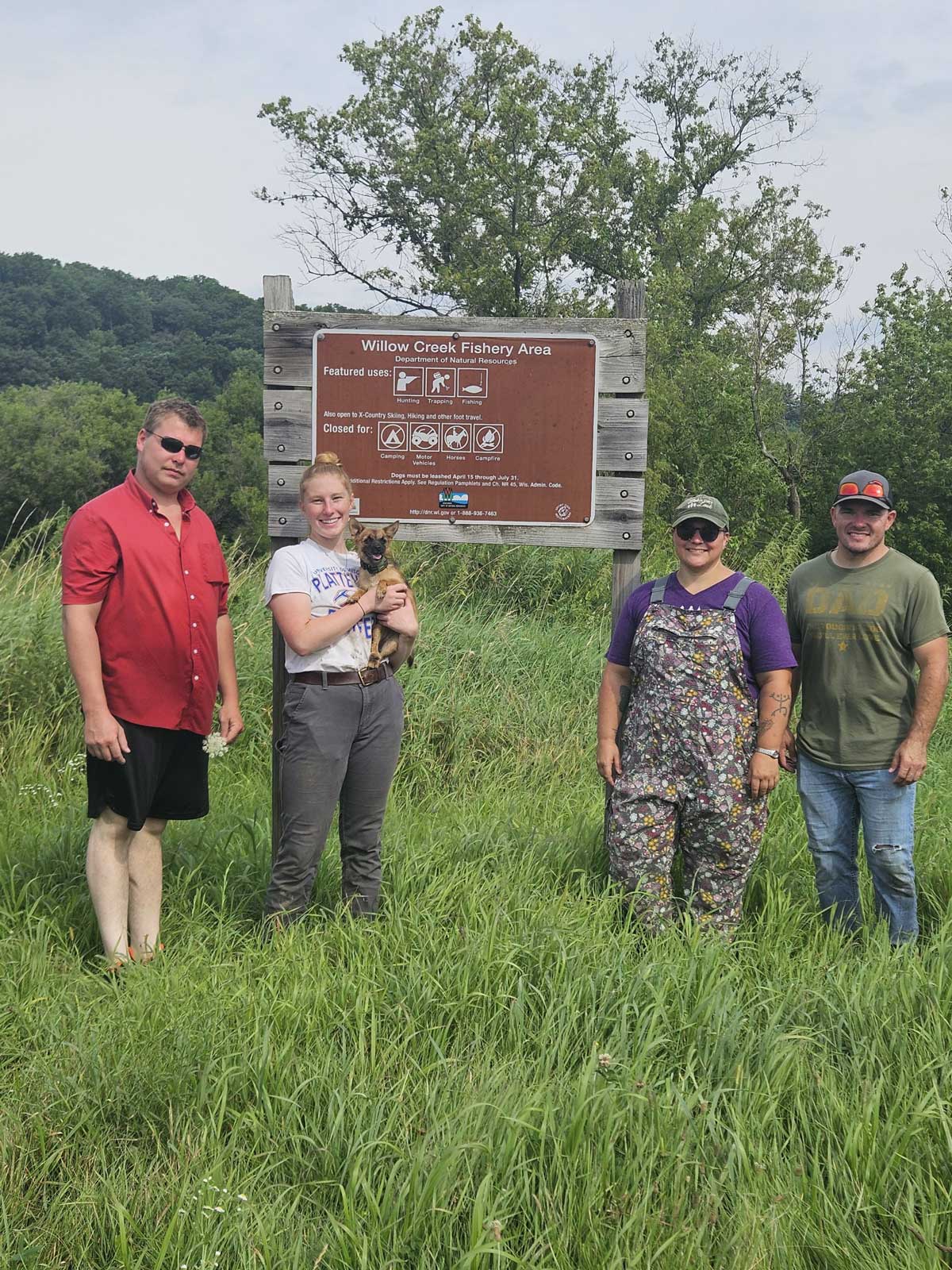 Four people standing in a field in front of a Willow Creek Fishery Area sign