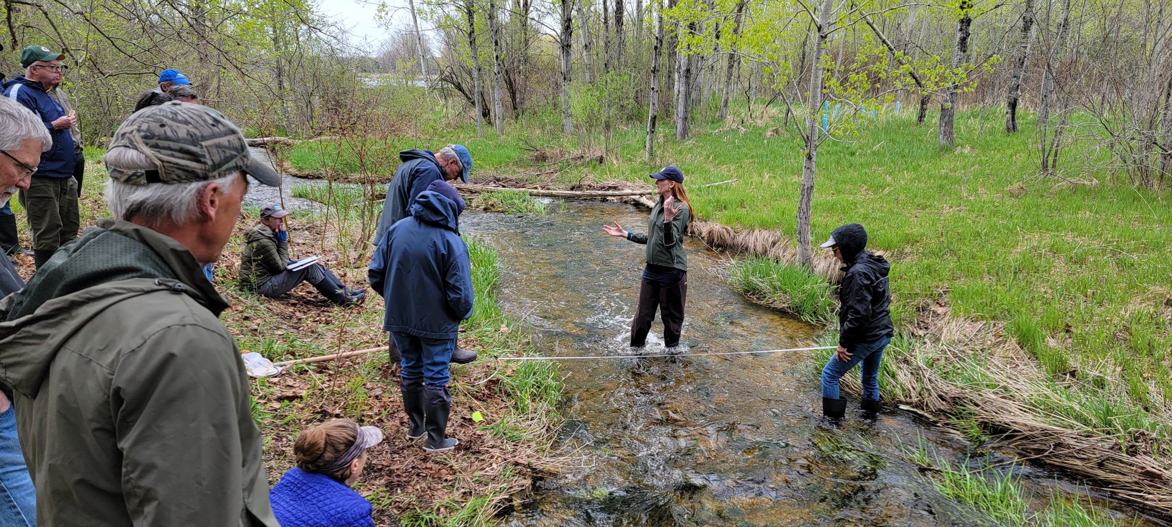 People stand on a river bank watching an instructor standing in the river. 