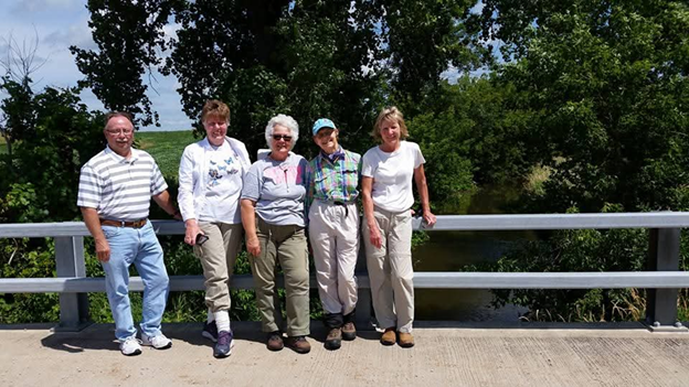 a group of five people stand on a bridge. In the background is a river and trees.