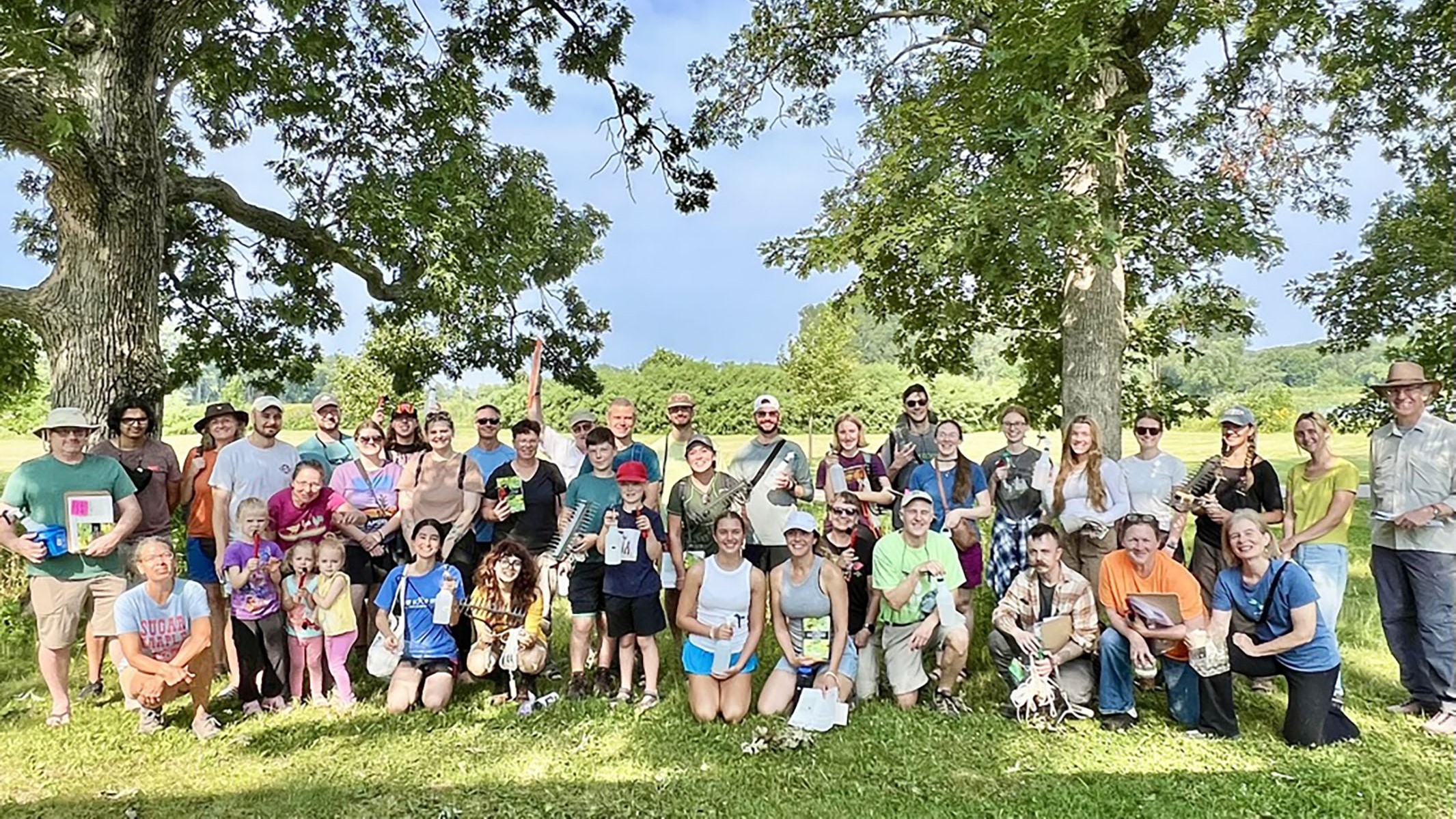 A large group of people pose for a photo. They are standing between trees outside on a sunny day
