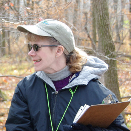 A woman in a beige baseball cap, sunglasses, and a blue windbreaker holds a clipboard. She is standing outside.