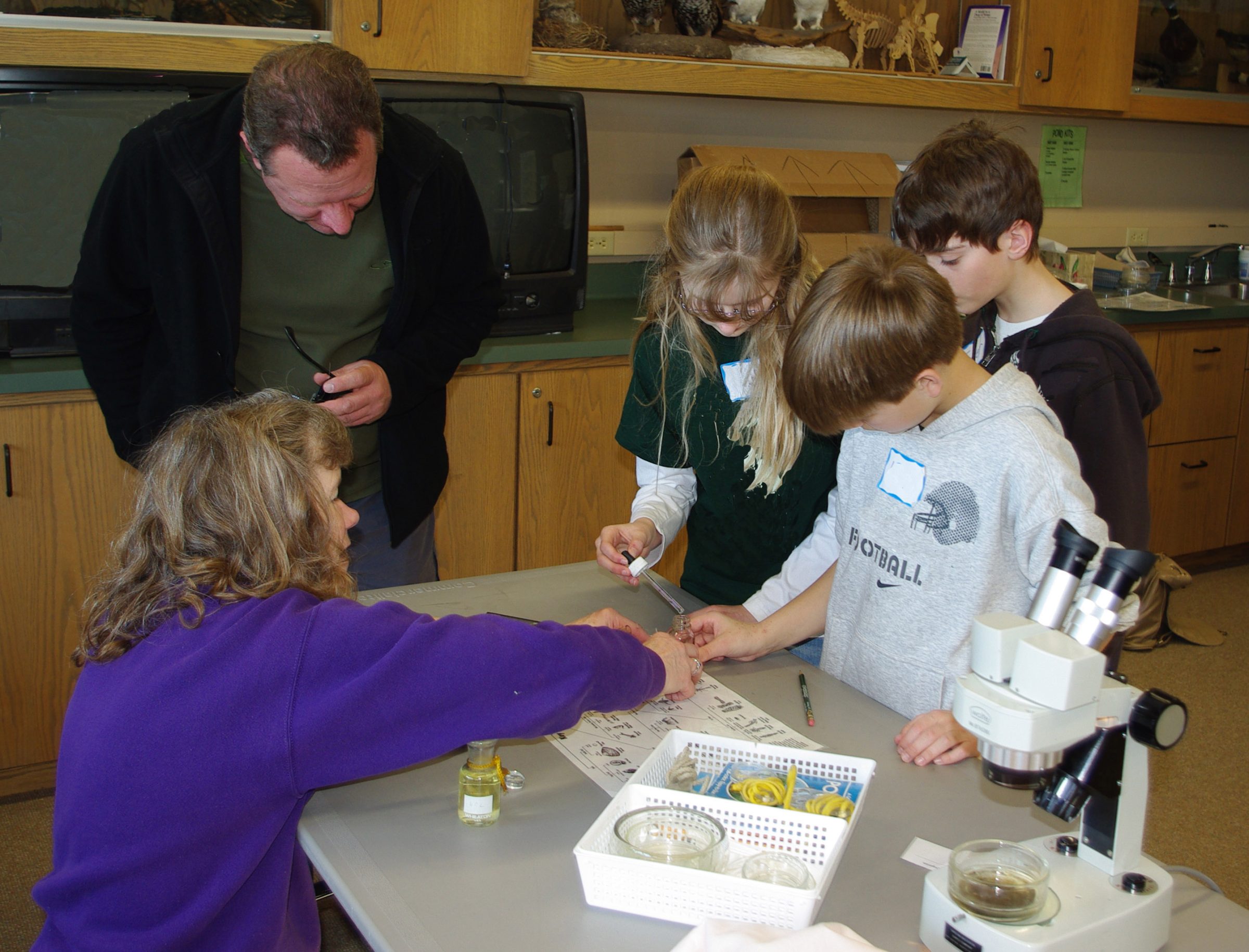 A woman in an indigo sweater teaches three young students about macroinvertebrates in a lab. 