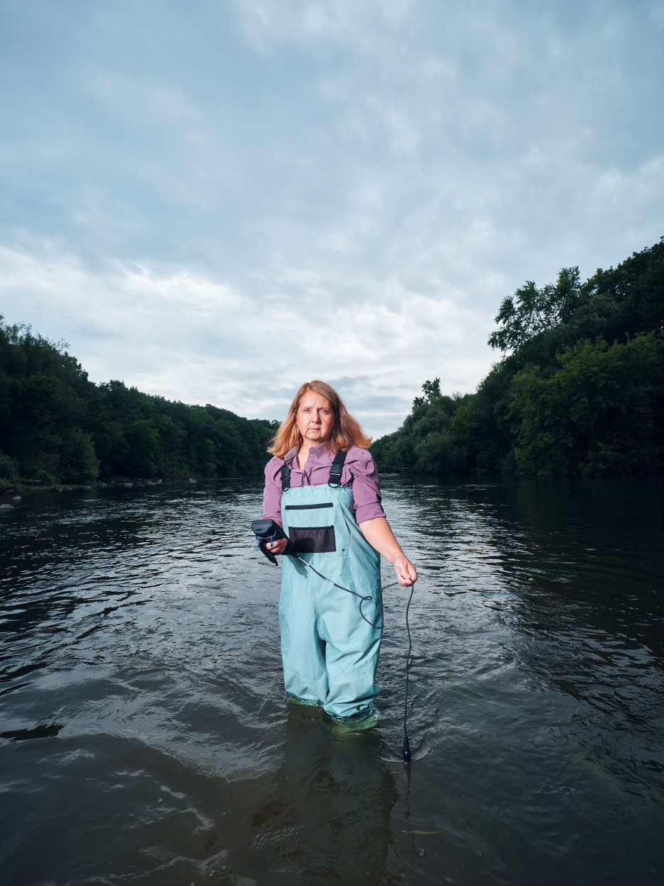 A blonde woman in blue bibs stands in a river holding monitoring equipment. It is an overcast day. 