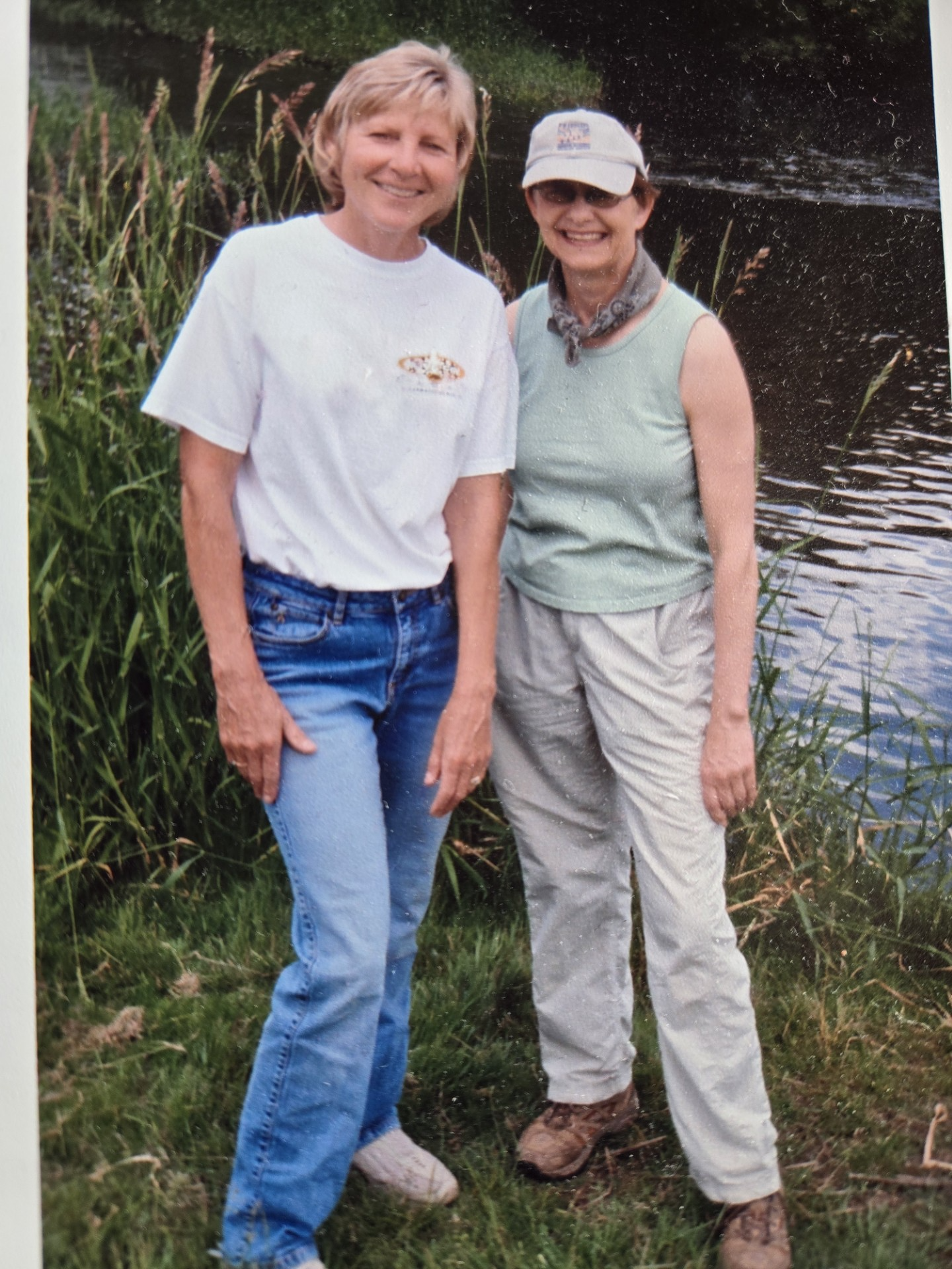 two woman stand next to a river smiling. The woman on the left is wearing jeans and a white t-shirt. The woman on the right is wearing a sage tank top and beige khakis. 
