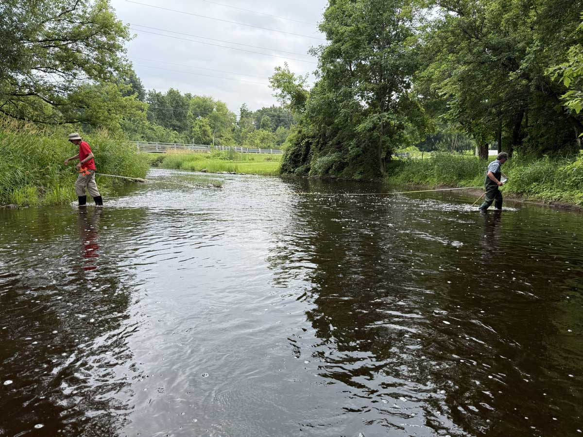 Two men walk across a river holding a net and measuring tape. They are measuring the width of the river. 