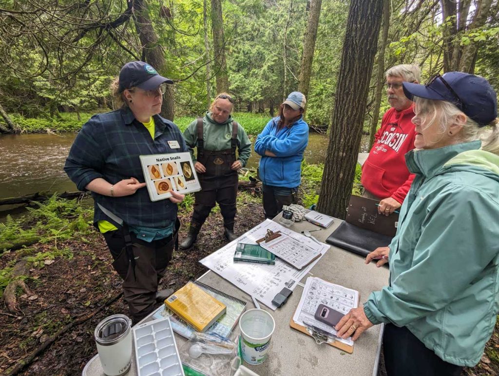 A group of people gather next to a wooded stream and learn from a presenter about native snails.
