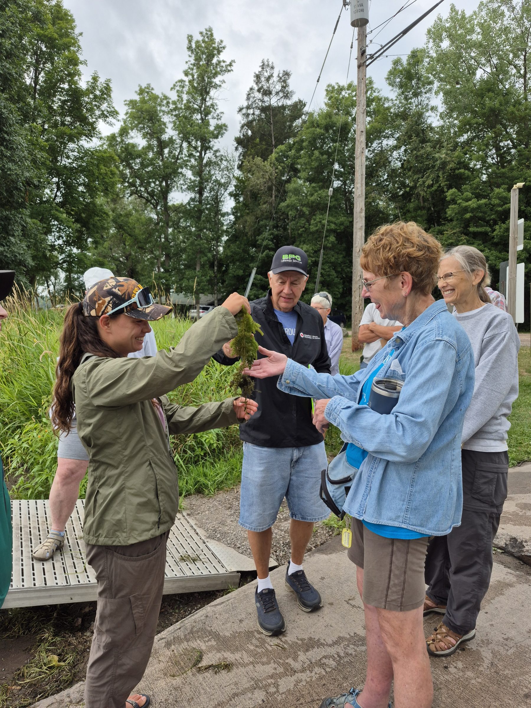 A group of people stand on a sidewalk. The two people in the foreground are standing facing each other holding a green AIS in their hands. The people in the background stand around them looking at the AIS. The background is green foliage.