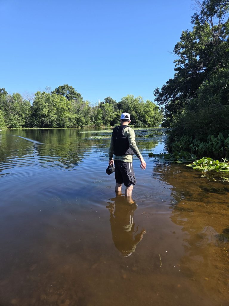 A man in black shorts and green long sleeve stands in brownish river water on a clear blue sunny day. His back is to the camera.
