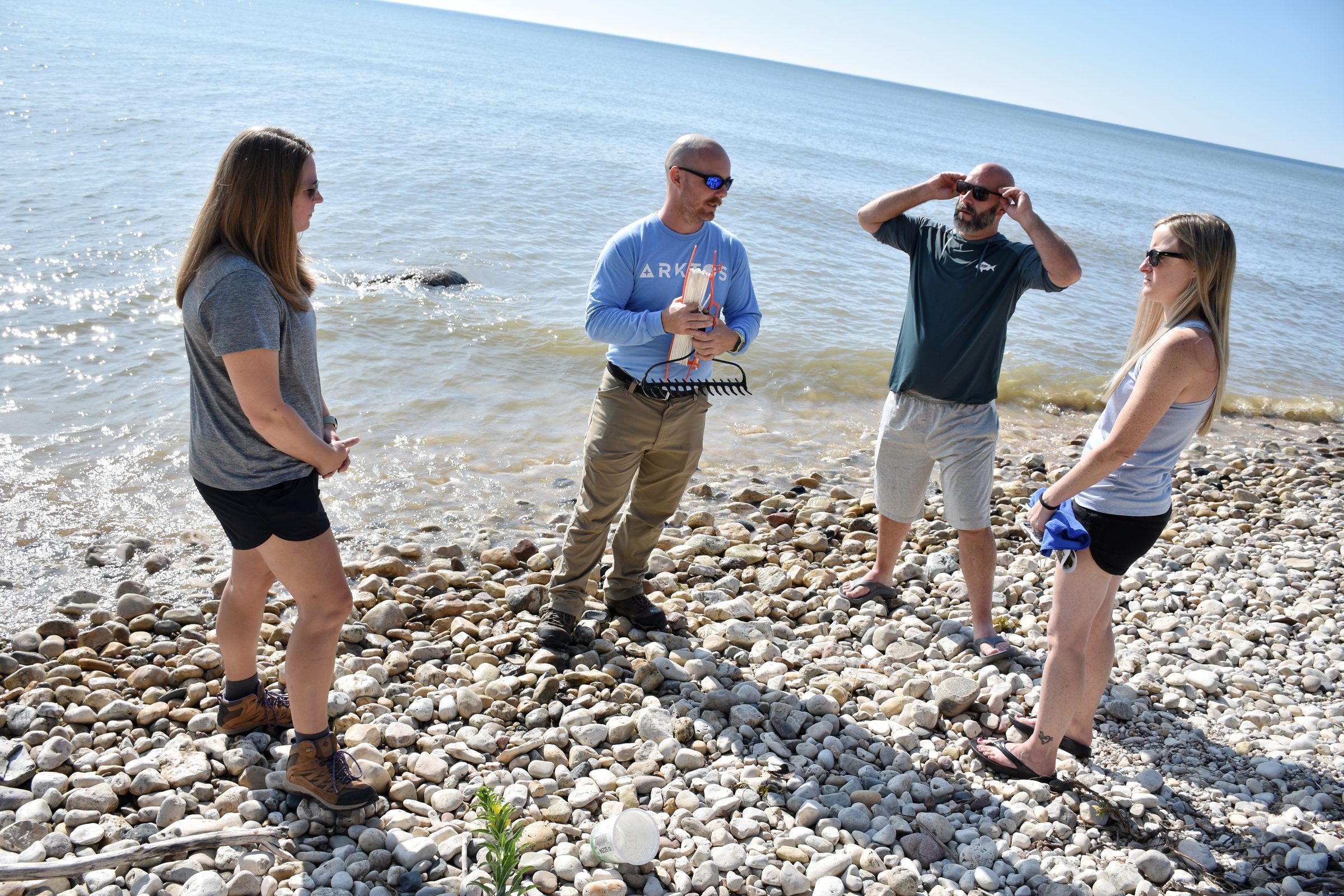 A man in a long sleeve baby blue shirt wearing sunglasses holds a rake head with white rope attached to it. The rope is coiled around an orange roll. A man with a dark grey shirt and khaki shorts stands next to him. The two people are standing on the rocky banks of a lake. The lake is behind them. It is sunny.