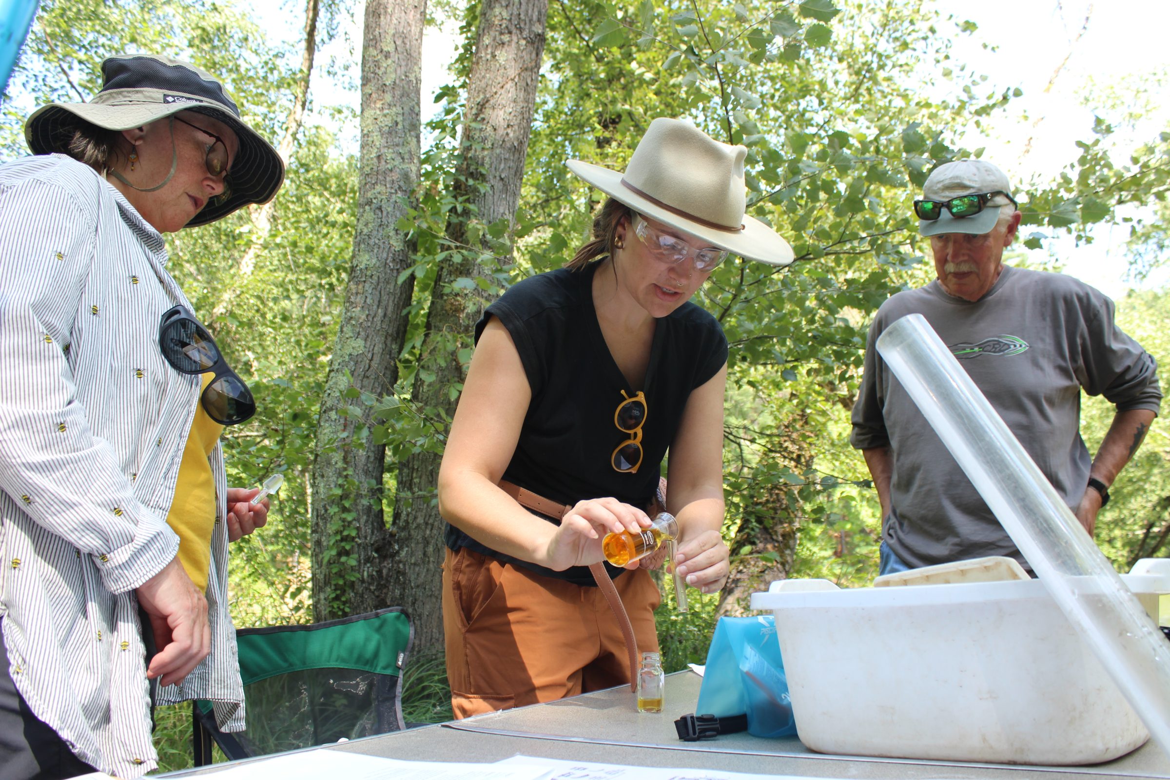 Three people stand around a foldout table with a white tray. They are measuring dissolved oxygen using a Hach Kit. The group is standing outside with trees behind them.