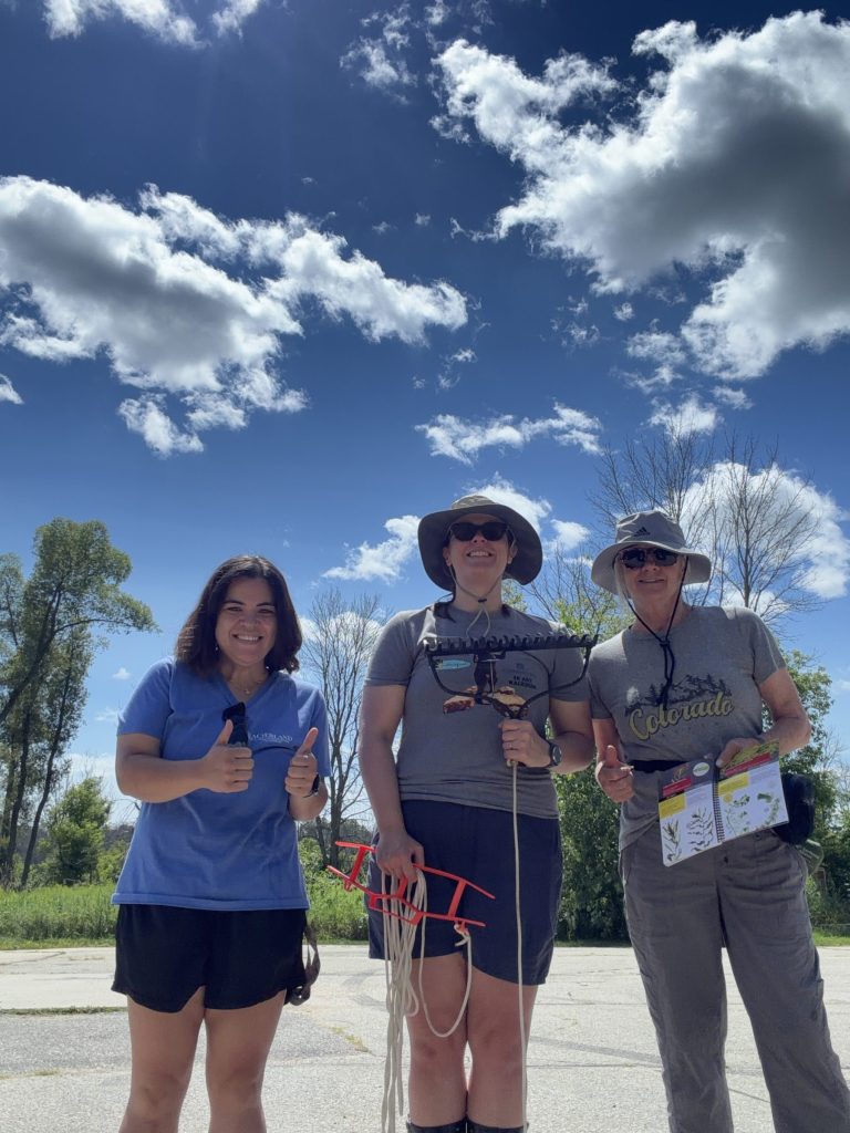 Three people stand posing for a photo. The person on the left has their hands in two thumbs up. The person in the middle is holding a rake head in one hand and a spool of rope in the other. The rope is attached to the rake head. The person on the right has one thumb up while the other hand holds open a field guide.
The sky is a dark blue with grey/white clouds.