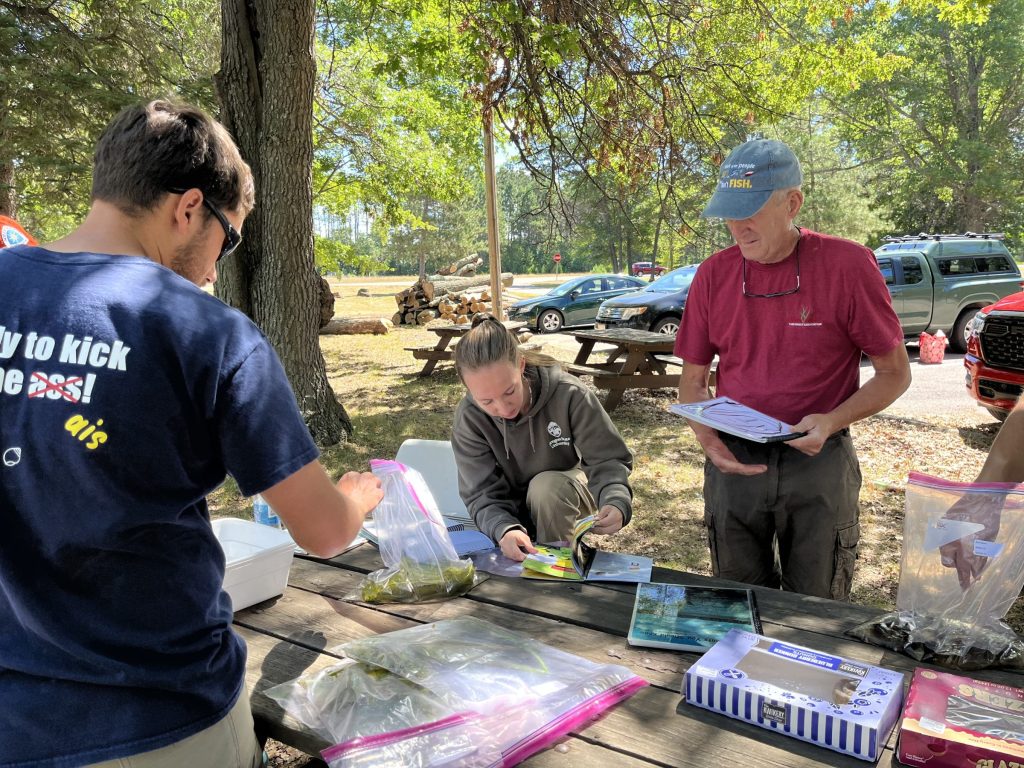 Three people stand around a wooden picnic table. They are looking at field guides. Ziploc bags are on the table. Two parked cars are in the background.