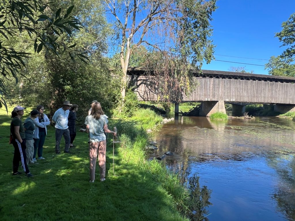 People stand on the left hand side of a river. The bank they are standing on is grassy and shaded. A bridge cross the river.