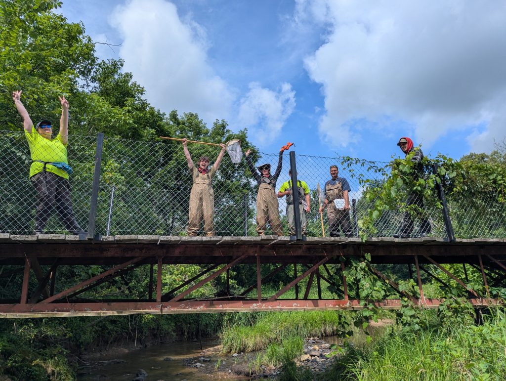 A group of five people stand on a bridge during a sunny day. Behind them are trees and clouds in the blue sky. Below the bridge is a stream. The group has their hands up in celebration. 