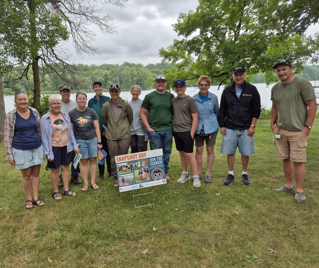 A large group of peope pose fo a photo in front of a small yard sign. They are standing on grass. In the background you can see a river with trees on the bank and a cloudy sky.