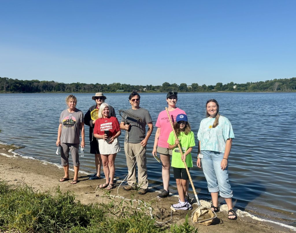 A group of people face towards the sun, posing for the photo. They are standing on a sandy shore of a lake. The lake is behind them.