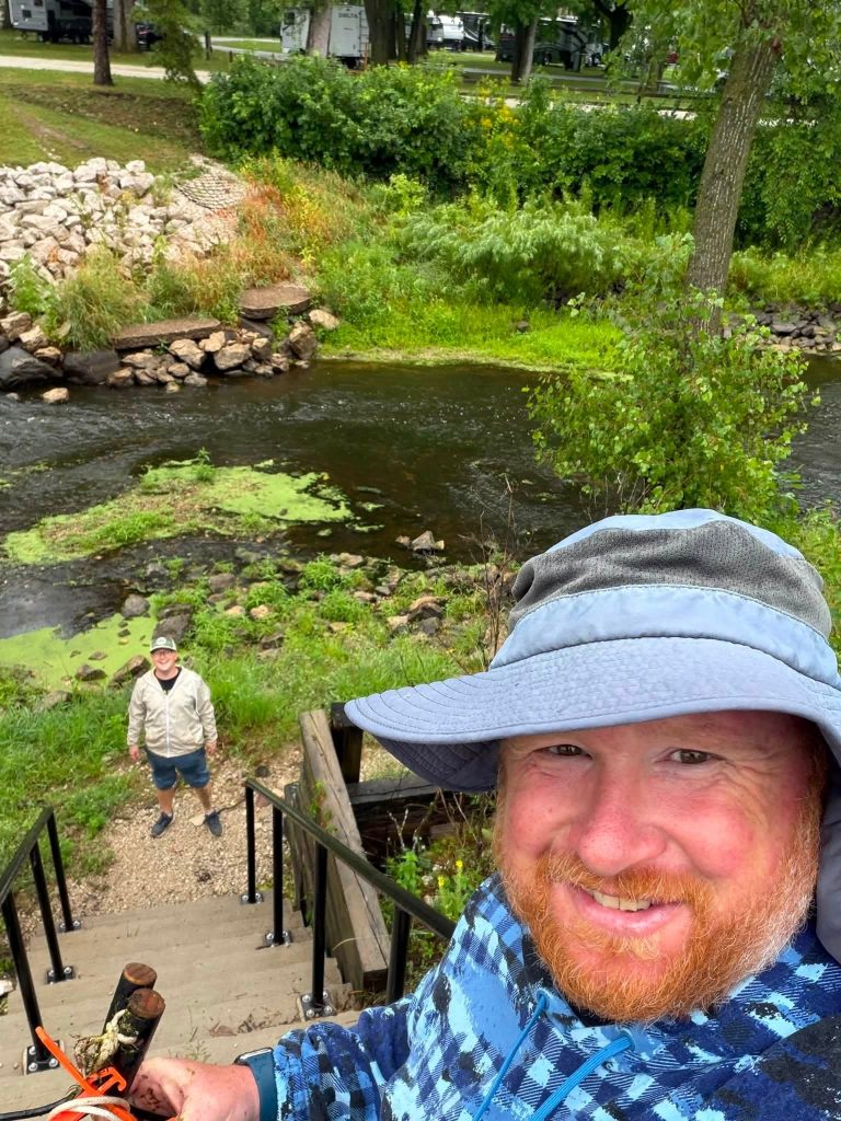 In the lower right hand corner is a close up of a man with a red beard wearing a blue bucket hat and blue long sleeves. He is taking a selfie, smiling at the camera. You can see he is at the top of some stairs.
At the bottom of the stairs is a person in a white jacket and blue shorts looking towards the camera, smiling. In the background is river with green foliage and rocks on the banks.