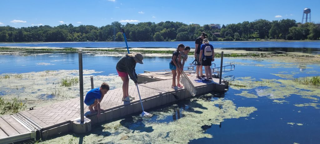 A group of kids stand on a floating plastic dock on a sunny day. They are holding nets in water that has green algae in it. In the background you can see the lake, trees, and a blue sky.